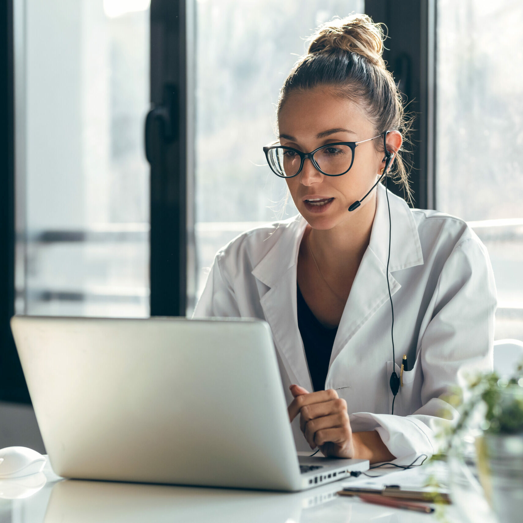 Shot of female doctor talking while explaining medical treatment to patient through a video call with laptop and earphones in the consultation.